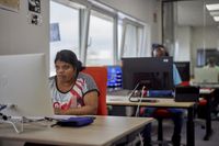 Werkruimte met 3 bureau's op een rij. Op elk bureau staat een computer. Twee mensen zitten aan een bureau, waarvan iemand een koptelefoon opheeft.