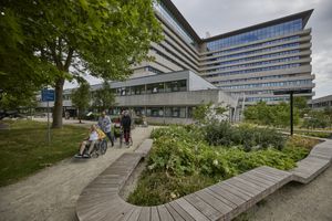 een groot modern gebouw met 10 verdiepingen, er wandelt een groepje mensen door de tuin.