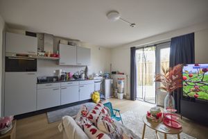 Open woonkamer en kitchenette met grijze kasten, bank met kussens met hartjespatroon, tv en tuindeur naar buiten.