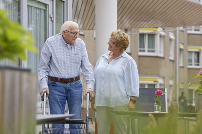 Een oudere man met rollator en vrouw met kort haar kijken elkaar aan. Ze staan buiten voor een appartementengebouw.