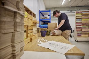 Een man staat aan een grote houten tafel en pakt iets in bubbelplastic. Naast hem staat een kartonnen doos op tafel en 2 bloempotten. Op de voorgrond ligt een grote stapel karton en een stoffen tas met opdruk van "Bijzonder Amsterdams".