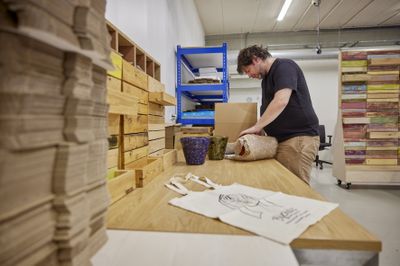 Een man staat aan een grote houten tafel en pakt iets in bubbelplastic. Naast hem staat een kartonnen doos op tafel en 2 bloempotten. Op de voorgrond ligt een grote stapel karton en een stoffen tas met opdruk van "Bijzonder Amsterdams".