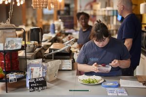 Een medewerkster met donkerblauw Bijzonder Amsterdams shirt schept een salade op een bord op de bar van het café.