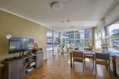 Moderne woonkamer met houten vloer, tv, eettafel en grote ramen met uitzicht over de stad. Kamerplanten maken de inrichting compleet.