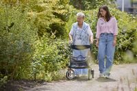 Een oudere dame loopt met een jonge medewerkster buiten met haar rollator. De zon schijnt, ze kijken naar de struiken.