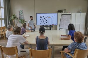 Een man met donker haar en een baard staat naast een groot beeldscherm. Daarvoor staat een grote, vierkante tafel en zitten er 6 mensen mee te kijken naar het scherm.