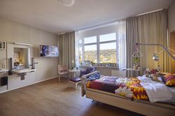 Kamer met een groot raam, bed, beddengoed met bloemenprint, tv aan de muur en stoelen; uitzicht op bomen.