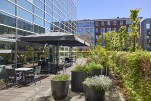 Een terras met tafels en stoelen onder parasols, omgeven door potplanten en groen, grenzend aan een glazen gebouw op een zonnige dag.