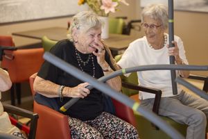 Twee oudere dames lachen tijdens het deelnemen aan een beweegactiviteit. Ze zitten op een stoel met een stok in hun handen.