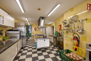 Een grote keuken met roestvrijstalen apparaten, een geblokte vloer, gele tegelwanden, hangend keukengerei en een krat met aardappelen op de grond.