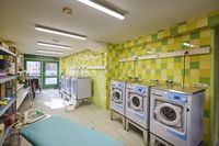 Lichte wasruimte met geelgroene tegelwanden, drie wasmachines, een strijkplank, planken met benodigdheden en een deur naar buiten.