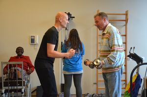 Een deelnemer wordt door een kale sportcoach aangemoedigd tijdens het heffen van gewichten. op de achtergrond zijn ook nog twee vrouwelijke deelnemers aan het sporten.