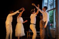 Vijf mensen met witte kleding staan in een donkere ruimte. Een van hen houdt een bal omhoog en de anderen reiken ernaartoe.