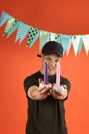 Een portretfoto van een jongen met pet met twee kandelaren met kaarsen in zijn hand. De achtergrond is kleurrijk met vlaggetjes.