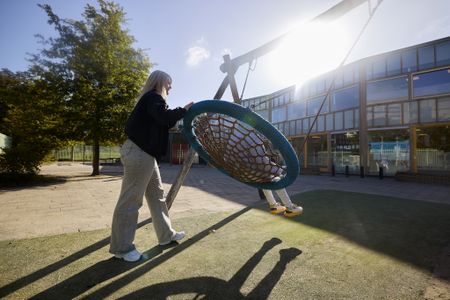 Op een zonnige dag duwt een vrouw een ronde schommel met daarin een kind op een speelplaats.
