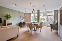 Lichte, moderne woonkamer met lichte meubels, een groene muur met bloemen erop, grote ramen en een houten vloer.