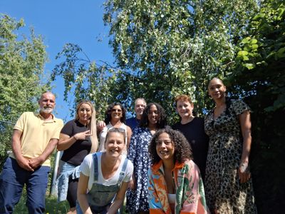 acht personen in kleurrijke en zomerse kleren kijken met open blik in de camera. De zon schijnt, op de achtergrond een boom vol in blad.