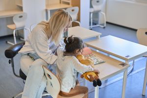 Een vrouw met blond haar zit naast een kindje aan een tafeltje in een klaslokaal. Ze lezen samen een kinderboek.