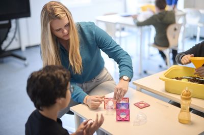 Een vrouw met lang blond haar en een kind zitten samen aan een tafeltje. Zij helpt hem met een puzzel.