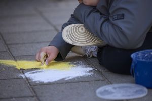 Een kind zit in kleermakerszit en tekent met geel krijt op de stoep, gekleed in een grijze jas.