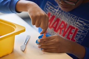Een kind in een superhelden-T-shirt boetseert klei met een lepel aan een tafel. Een gele bak staat ernaast.
