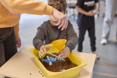 Een kind speelt met bonen en een trechter in een gele bak. Een volwassene helpt mee.