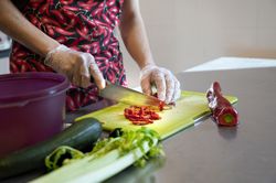 Persoon met schort aan met rode pepers, is met latex handschoenen aan zoete paprika's aan het snijden op een snijplank. Er ligt ook een courgette en een struik bleekselderij bij