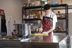 Persoon met schort aan met rode pepers is groente aan het snijden op een snijplank staande aan een keukenblok. Er staat een grote pan op het fornuis