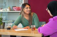 Een jonge vrouw met lang bruin haar en een groene trui zit met een pen in de hand aan tafel. Zij kijkt vriendelijk en met interesse naar een dame met fuchsia blouse en zwarte hoofddoek. Op tafel staat een kop thee en een kop koffie.