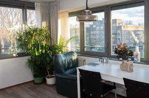 Een licht appartement op de hoek van het gebouw. De zon schijnt naar binnen op de grote planten. Er staan een eettafel met 2 stoelen en een fauteuil.