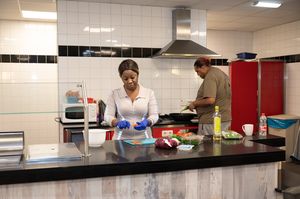 Twee mensen bereiden een maaltijd in een grote keuken. Er staan ingrediënten op het aanrecht.