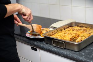 Persoon schept lasagne uit metalen ovenschaal op een bord met een lepel en een houten spatel