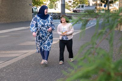 Een stevige dame met donkerblauwe hoofddoek loopt buiten met stevige pas en kijkt lachend naar een client naast haar met halflang bruin haar met een lach op haar gezicht.