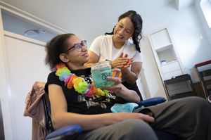 Een djonge vrouw met bril en zwart krullend haar zit in een rolstoel in de badkamer en houdt een pot blauwe haargel in haar hand. Een dame met zwarte vlechten staat naast haar en wrijft gel in haar handen. Ze kijken lachend naar elkaar