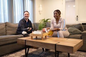 Twee lachende personen zitten op een comfortabele hoekbank. De één heeft een afstandsbediening van en de ander een tijdschrift.