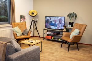 In een gezellige tv.hoek van de gemeenschappelijke ruimte staat een grijzde bank, twee bruin leren fauteuils en een televisie op een modern tv meubel.