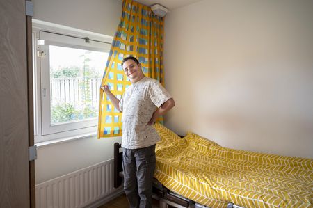 Jonge man staat in slaapkamer voor een bed met gele print en houdt het gordijn vast dat geel is met blauwe blokjesprint.