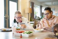 Een jonge man en vrouw met allebei een bril zitten te eten aan tafel in een woonkamer. Ze eten brood en soep en op tafel staan ook bakjes met komkommer en tomaat.