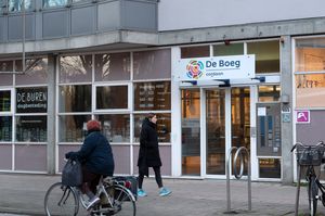 Ingang met daarboven een bord met het Cordaan logo en "De Boeg" in blauwe letters. Er loopt iemand voorbij op de stoep en er fietst iemand voorbij op het fietspad. Rechts van de entree staat "De Buren Dagbesteding" en de openingstijden in witte letters.