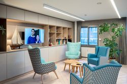 Woonkamer met links een kastenwand met tv. Er staan 4 blauwe fauteuils, een grote plant en 3 bijzettafeltjes.