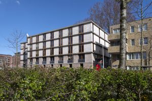 Achter een groene heg staat een grijs gebouw met vijf verdiepingen en bruine kozijnen.