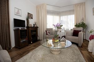 In een kamer met twee grote ramen staan twee lichte leunstoelen met ervoor een glazen salontafel. Aan de muur hangen foto's van een jong stel en een baby.