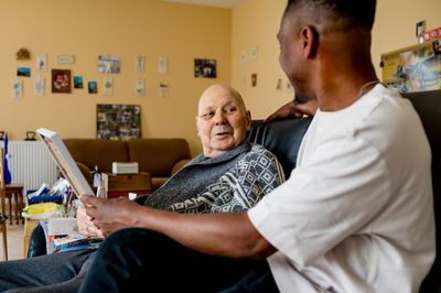 Een jonge man met donker haar en wit shirt zit naast een oudere, kale man in een leren stoel. Ze houden samen een fotolijst vast en praten met elkaar.