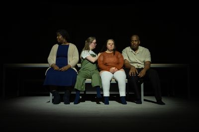 Een donkere foto met vier mensen op een bankje op het toneel. Ze kijken verdwaasd.