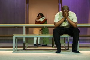 Een man zit met zijn handen voor zijn gezicht op een bankje op het toneel. Achter hem omhelzen twee mensen elkaar.