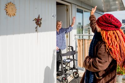 Thuiszorgmedewerker zwaait naar oudere dame die met rollator in deuropening van woning staat