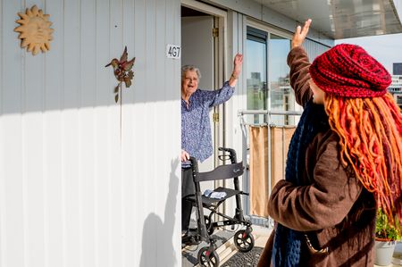 Thuiszorgmedewerker zwaait naar oudere dame die met rollator in deuropening van woning staat