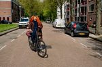 Een vrolijke dame met oranje dreadlocks fietst lachend over een straat met flats.