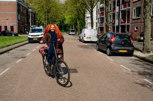 Een vrolijke dame met oranje dreadlocks fietst lachend over een straat met flats.