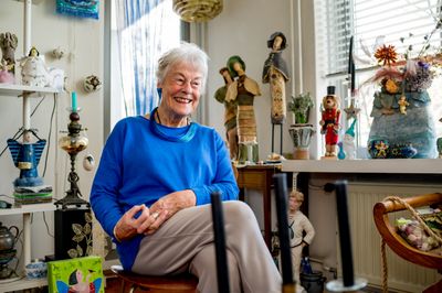 Oudere dame met blauwe trui en blauwe accessoires zit in een kamer vol kunstige beelden en lampen en kijkt blij opzij